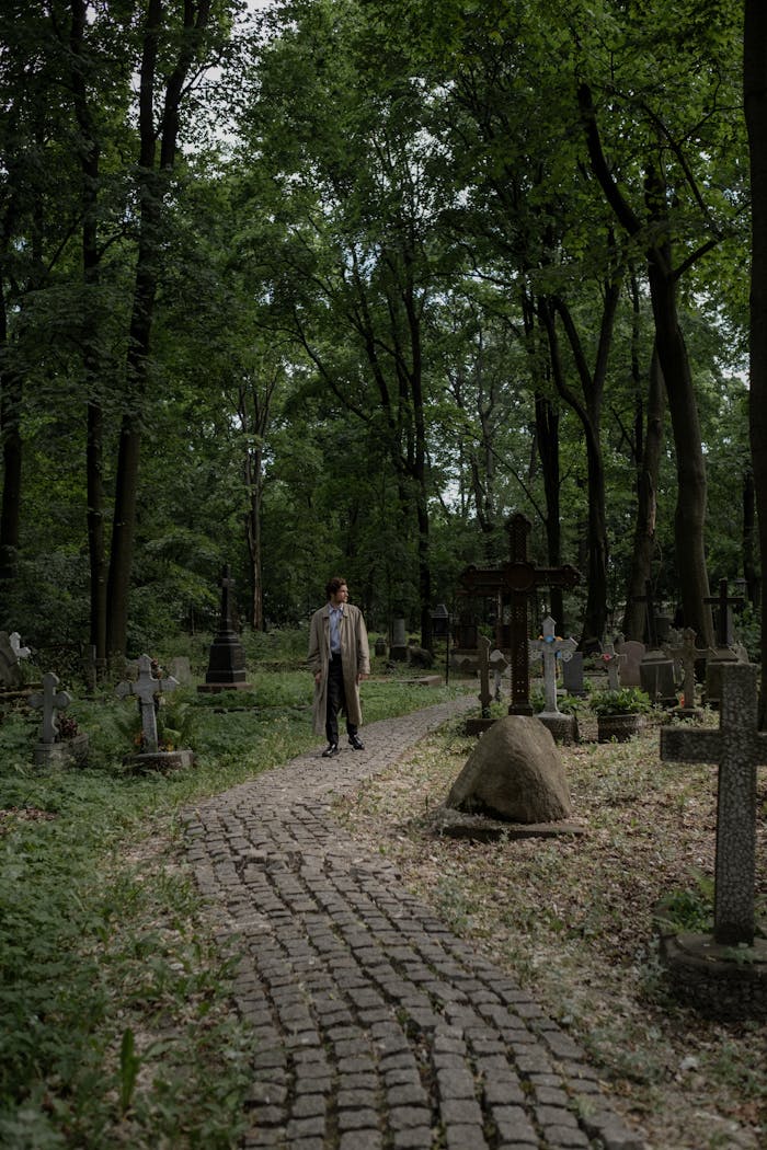 services-02 A person stands alone on a cobblestone path in a lush, tranquil cemetery.